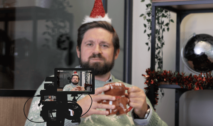 A man wearing a Santa hat and Christmas jumper, holding a mug shaped like a gingerbread man, posing in front of a camera